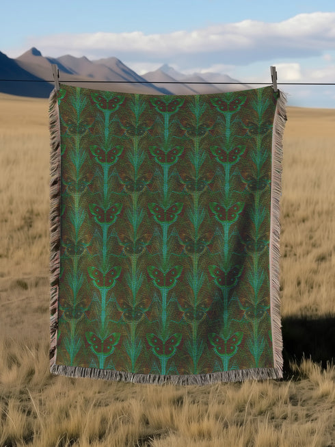 A woven cotton throw featuring an intricate luna moth design in deep green and russet tones, framed by soft fringe.